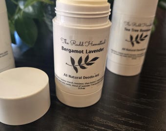 May include: A white cylindrical deodorant stick with a label reading The Rudd Homestead Bergamot Lavender All Natural Deodorant 2.5 oz. The container is open, revealing the solid white product inside, with a matching white cap resting nearby.