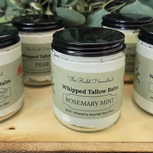 May include: Several glass jars of whipped tallow balm with black lids are arranged on a wooden tray. Each jar has a label with the text "The Rudd Homestead" and the balm's scent, including "Signature Blend," "Rosemary Mint," and "Vanilla."