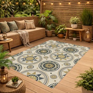 May include: An outdoor rug with a cream background and a repeating pattern of navy blue and gold circles. The circles have starburst and dot designs. The rug is on a wooden deck, with outdoor furniture and plants in the background.