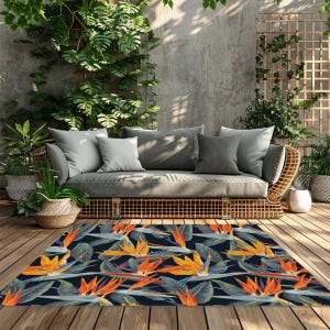 May include: Outdoor rug with a vibrant floral pattern featuring orange and yellow bird-of-paradise flowers on a dark blue background. The rug is placed on a wooden deck in front of a wicker sofa with gray cushions, creating a tropical outdoor living space.