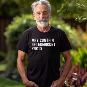 May include: A man wearing a black t-shirt with the text "MAY CONTAIN AFTERMARKET PARTS" in white lettering. He is standing in front of a green background.