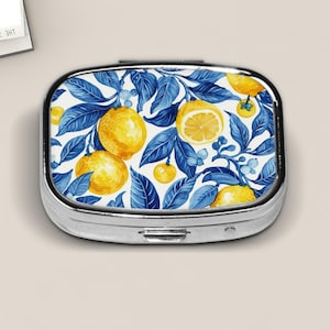 May include: A silver pill box with a watercolor design of lemons, blue leaves, and small berries on a white background. The box is rectangular with rounded edges and a hinged lid. The design features bright yellow lemons and deep blue leaves.