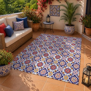 May include: A rectangular area rug featuring a repeating floral tile pattern in shades of blue, red, and yellow. The rug is laid out on a terracotta tiled patio floor, positioned in front of a wicker sofa with blue and red throw pillows.