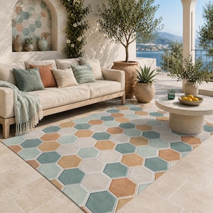 May include: A rectangular area rug featuring a repeating geometric pattern of interlocking hexagons in muted shades of sage green, terracotta orange, and cream. The rug is laid flat on a light-colored stone patio floor in an outdoor living space.