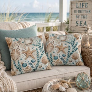 May include: Two square throw pillows featuring a coastal pattern of tan seashells, beige starfish, and teal seaweed on a light background. The pillows are placed on a light-colored sofa, with a sign reading LIFE IS BETTER BY THE SEA nearby.