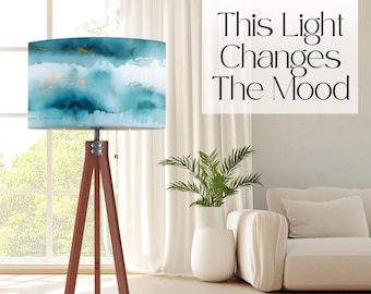 May include: A floor lamp with a tripod base made of dark wood and a cylindrical lampshade featuring an abstract watercolor pattern in shades of teal, blue, and white with gold accents. The text "This Light Changes The Mood" is visible.
