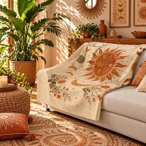 May include: A cream-colored throw blanket featuring a large, central sun face illustration in shades of orange and brown, surrounded by crescent moons, stars, and floral vine motifs. The blanket is draped over a sofa, showcasing a detailed celestial pattern.