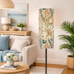 May include: A tall cylindrical floor lamp shade featuring a beige background with a repeating pattern of brown starfish, various seashells, teal seaweed, and small circular bubbles. The shade is supported by a thin, dark, vertical metal pole stand.