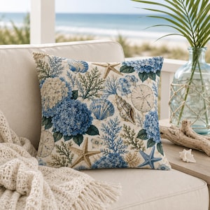May include: A square throw pillow featuring a detailed print of blue hydrangea flowers, various seashells, starfish, and coral branches on a light beige background. The design incorporates shades of deep blue, light blue, and tan throughout.