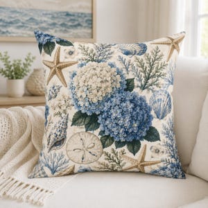 Könnte beinhalten: Ein quadratisches Dekokissen mit einem Küsten-Motiv auf cremefarbenem Hintergrund. Das Design zeigt blaue und weiße Hortensienblüten, durchsetzt mit beigen Seesternen, verschiedenen Muscheln, Sanddollars und blauen Korallenzweigen.