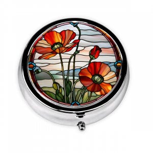 May include: A round silver pill box with a stained glass design of red poppies on a blue and white background.