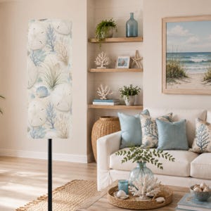 May include: A tall floor lamp featuring a cylindrical shade decorated with a pattern of white sand dollars, blue coral, and green sea grass. The lamp has a simple black metal pole stand and is positioned in a room with coastal decor elements.