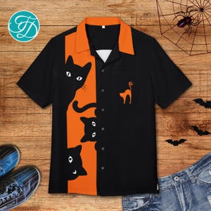 May include: Black button-up shirt with orange accents and a cat-themed design. The shirt features a black cat graphic on the front and a small orange cat on the pocket. The collar and center panel are orange.