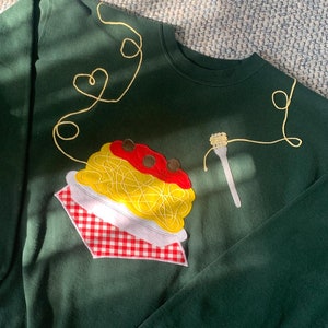 May include: A dark green sweatshirt with a yellow, red, and white embroidered spaghetti and meatballs design. The spaghetti is tied in a heart shape on the left side of the sweatshirt.