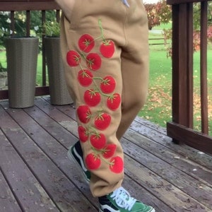 May include: A woman wearing a green crop top and brown sweatpants with a red tomato vine embroidered on the left leg. She is wearing sunglasses and cherry earrings. She is standing on a wooden deck.