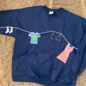 Puede incluir: Una sudadera azul marino con una línea blanca y varias prendas colgadas en ella, incluyendo calcetines, una camiseta, un sujetador y un vestido.
