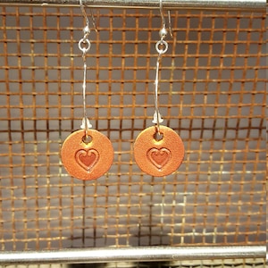 Op de afbeelding: Een paar oorringen met een zilveren ring en een ronde, oranje leren hanger met een hartvormig ontwerp.