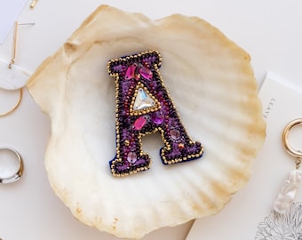 May include: A purple and pink beaded letter A brooch in a white seashell. The brooch is surrounded by other jewelry, including a gold chain necklace, a pair of gold hoop earrings, and a pair of pearl earrings.