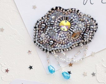 Può includere: Un broche a forma di nuvola decorato con perline, con un grande cristallo iridescente al centro. Il broche è composto da perline argentate e blu e da una frangia di perline argentate con due perline blu a forma di lacrima che pendono. Il broche è su uno sfondo bianco con stelle argentate.