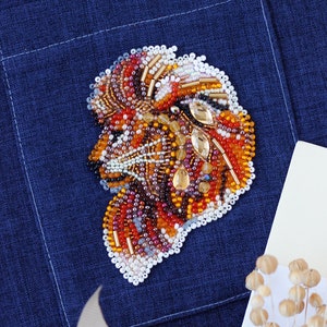 Può includere: Un'applicazione di testa di leone in perline colorate di arancione, marrone e bianco, cucita su una tasca in tessuto denim blu scuro.