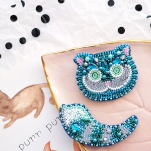 May include: Two blue and green beaded cat brooches on a pink and gold dish. The cat brooches are in the shape of a cat's head and a cat's tail. The text "purr purr" is visible in the background.