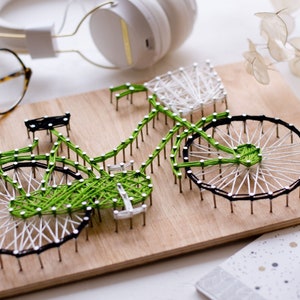 May include: Green and white string art of a bicycle on a wooden board. The bicycle has a basket on the handlebars and a green frame.
