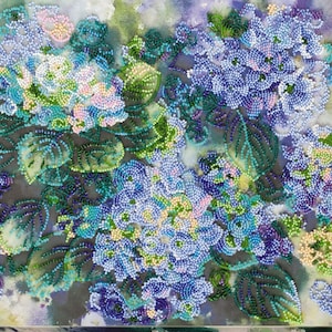 Puede incluir: Primer plano de una obra de arte con cuentas que representa un ramo de hortensias azules y moradas con hojas verdes. Las flores están dispuestas en un racimo y parecen estar hechas de pequeñas cuentas.
