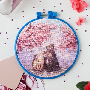 May include: A blue embroidery hoop with a cross-stitch design of two cats under a pink cherry blossom tree. The hoop is on a white surface with pink flowers and purple glitter stars.