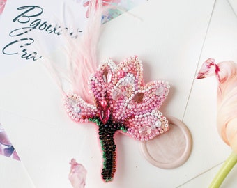 Può includere: Un broche a forma di fiore rosa perlato con uno stelo verde e perline marroni. Il broche è su una superficie bianca con una busta rosa e un fiore rosa.