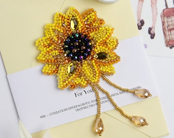 Può includere: Una spilla a forma di girasole realizzata con perline, con uno schema di colori giallo, arancione e oro. Il centro del girasole è un gruppo di perline viola scuro e nere. La spilla ha tre fili di perline pendenti con perline trasparenti e ambrate.