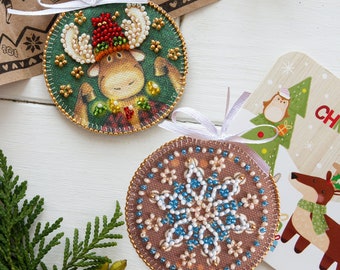 Puede incluir: Dos adornos navideños hechos a mano. Un adorno presenta un alce marrón con cuernos rojos y un gorro rojo sobre un fondo verde. El otro adorno presenta un copo de nieve azul y blanco sobre un fondo marrón.