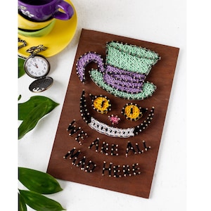 Puede incluir: Arte de hilo del Gato de Cheshire de Alicia en el País de las Maravillas. La cara del gato está hecha de hilo negro, con hilo blanco para la sonrisa y hilo amarillo para los ojos. El gato lleva un sombrero verde y morado. Las palabras "We are all mad here" están escritas debajo de la cara del gato.