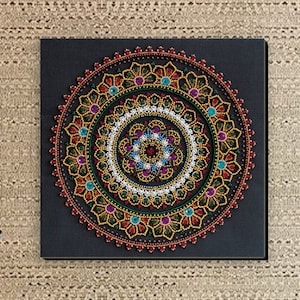 May include: A black canvas with a colorful mandala design made of beads. The mandala is surrounded by a black border and is set against a beige patterned wall.