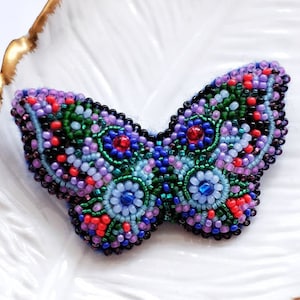 May include: A beaded butterfly brooch with a detailed design in shades of purple, blue, green, and red. The brooch is displayed on a white, feather-shaped dish with a gold rim. A small jar of gold glitter is in the foreground.