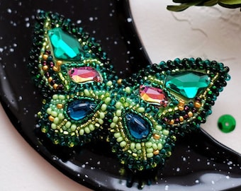 May include: A green and blue beaded butterfly brooch with iridescent accents on a black crescent moon shaped plate.