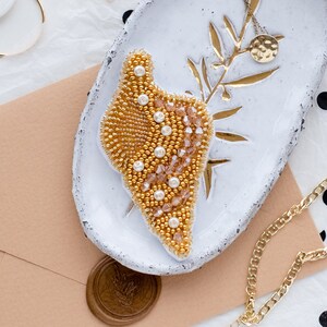 May include: A white ceramic dish with a gold beaded design, a gold chain necklace with a hammered gold pendant, and a gold chain bracelet with a white pearl charm. The dish is on a white surface with black polka dots.