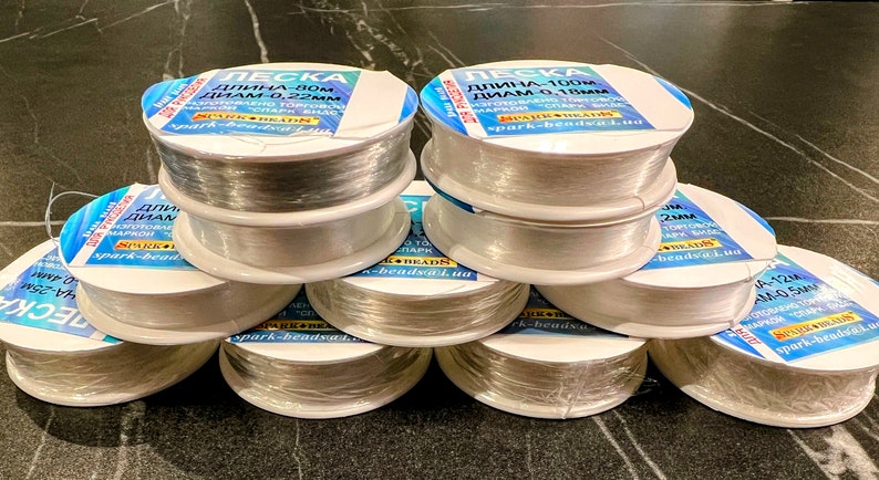 May include: A stack of clear spools of fishing line, each with a label in Russian. The labels indicate the length and diameter of the line. The spools are stacked in a pyramid shape on a dark surface.