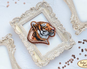 Könnte beinhalten: Eine Tigerkopf-Brosche aus Perlen in Braun-, Orange- und Weißtönen, präsentiert in einem weißen, verzierten Rahmen. Die Brosche ist eine detaillierte und realistische Darstellung eines Tigerkopfes.