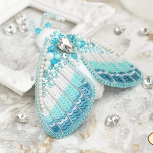 May include: A blue and white beaded moth brooch with a crystal accent. The brooch is handmade and features a delicate design.