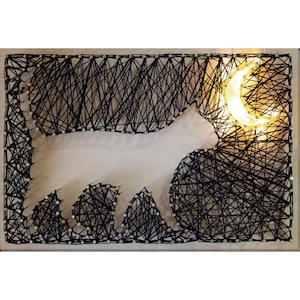 May include: A handmade string art piece featuring a white cat silhouette walking towards a glowing crescent moon. The cat is set against a backdrop of black string, creating a night sky effect. The artwork is mounted on a wooden board.