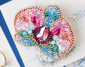 Può includere: Un broche di perline colorate con perline rosa, blu e verdi e strass. Il broche ha la forma di un fiore e ha un bordo dorato.