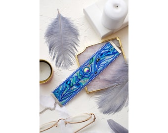 May include: A blue beaded bracelet with a floral design and iridescent accents. The bracelet is on a white surface with feathers and a candle.