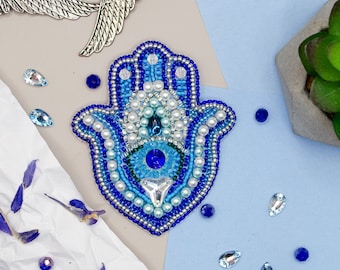 May include: A blue and white beaded hamsa hand with a blue evil eye in the centre. The hamsa is surrounded by silver beads and is on a white and blue background.