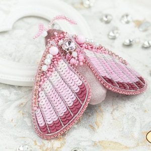 May include: A pink and white beaded moth brooch with a crystal embellishment. The brooch is handmade and features a delicate design.