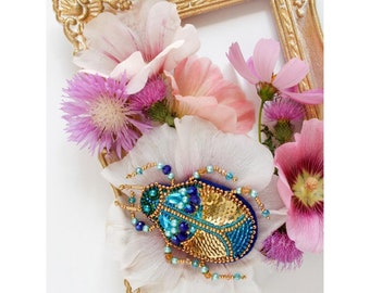 Può includere: Una spilla a forma di scarabeo blu e oro con accenti blu iridescenti. La spilla è circondata da fiori rosa e bianchi.