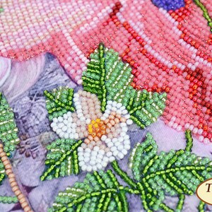 K&ouml;nnte beinhalten: Nahaufnahme eines Blumenmusters aus Perlen mit rosa, wei&szlig;en, gr&uuml;nen und orangefarbenen Perlen. Die Blume ist von gr&uuml;nen Bl&auml;ttern umgeben. Das Muster befindet sich auf einem Stoffhintergrund mit dem Text "Tela Artis" in der unteren rechten Ecke.