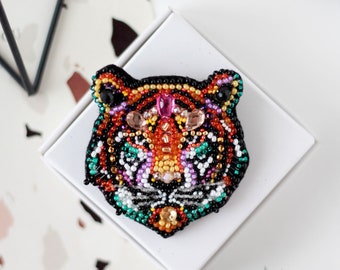 Puede incluir: Un broche de cabeza de tigre con cuentas de color naranja, negro, blanco, verde, rosa y dorado. El broche está sobre una superficie blanca.