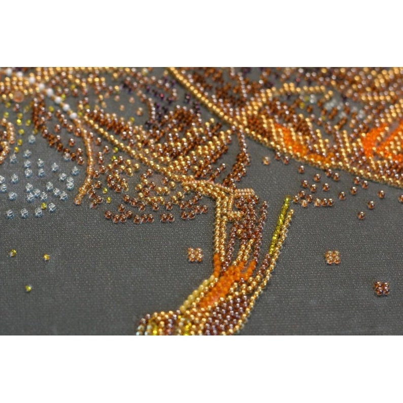May include: Close-up of a beaded design on a dark gray fabric. The beads are in shades of gold, brown, and orange, creating a swirling pattern.