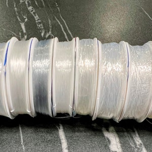 May include: A close-up of ten spools of clear fishing line, each wound around a white plastic spool with a blue stripe. The spools are arranged in a row, with the ends of the line visible.