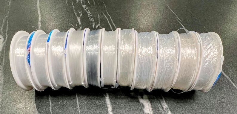 May include: A close-up of ten spools of clear fishing line, each wound around a white plastic spool with a blue stripe. The spools are arranged in a row, with the ends of the line visible.